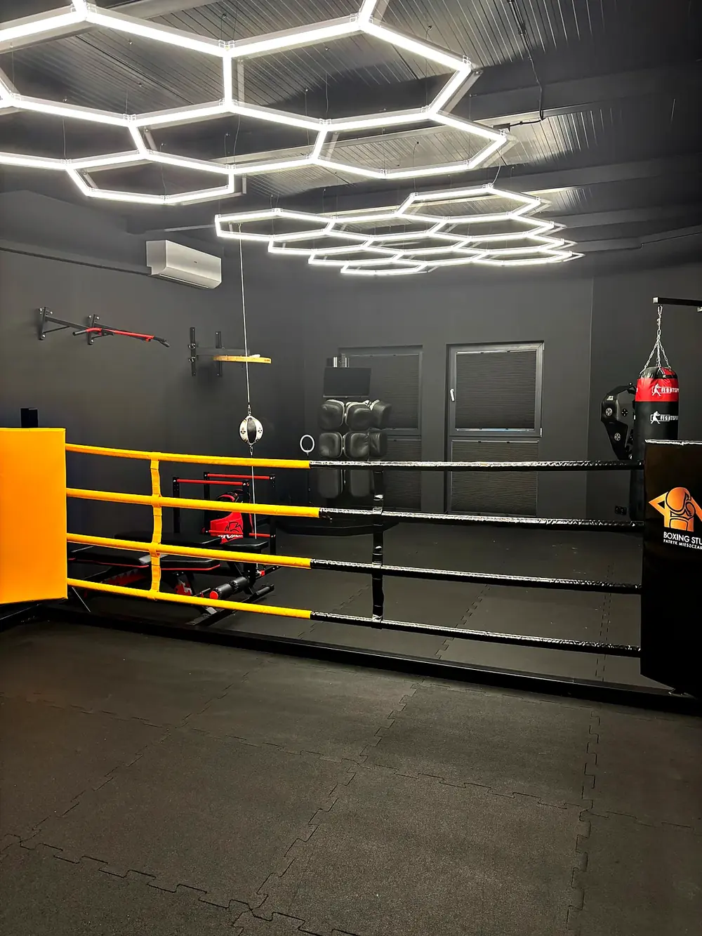 Ring treningowy i worki bokserskie w sali PM Boxing Studio pod heksagonalnym oświetleniem